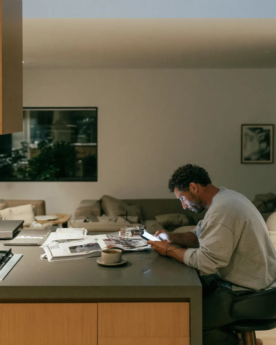 A man at a kitchen counter at first light, reading his phone over a cup of coffee.